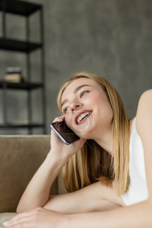Cheerful blonde woman talking on smartphone while lying on couch at home 