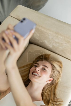 Top view of cheerful blonde woman taking selfie on blurred mobile phone at home 