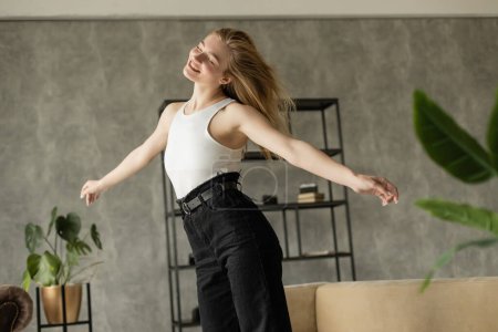 fröhliche blonde Frau tanzt mit geschlossenen Augen, während sie zu Hause Spaß hat