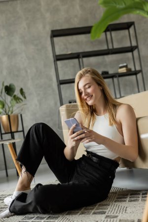 mujer rubia sonriente con teléfono inteligente sentado en la alfombra cerca del sofá en la sala de estar moderna
