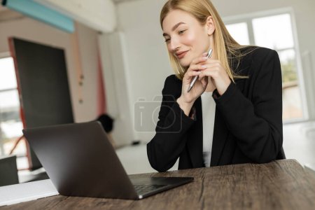 Photo for Smiling coach holding pen and looking at laptop during video call at home - Royalty Free Image
