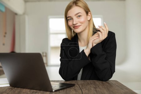 entrenador feliz en blazer negro mirando la cámara cerca de la computadora portátil en casa