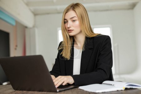 Blondine im schwarzen Blazer tippt während der Arbeit zu Hause auf Laptop neben Notizbuch