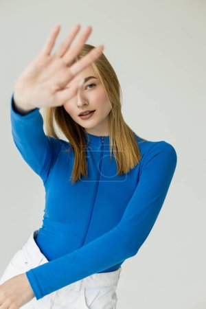 trendy woman in blue long sleeve shirt standing with outstretched hand on blurred foreground isolated on grey