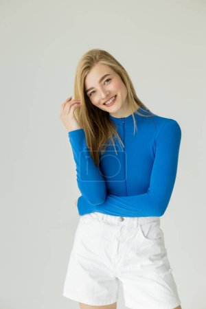 Photo for Cheerful blonde woman in blue turtleneck and white shorts looking at camera isolated on grey - Royalty Free Image