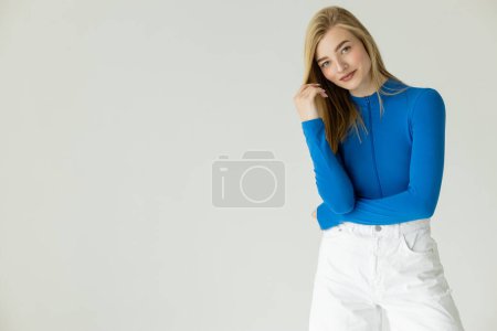 smiling blonde woman in blue turtleneck and white shorts looking at camera isolated on grey