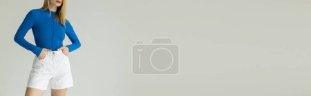Téléchargez les photos : Vue partielle de la femme en chemise bleue à manches longues debout avec les mains dans des poches de short blanc isolé sur gris, bannière - en image libre de droit