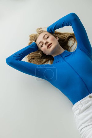 high angle view of blonde woman in blue long sleeve shirt lying with hands behind head on grey background 
