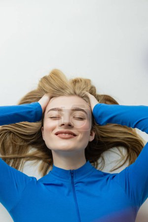 top view of young blonde woman smiling with closed eyes while lying on grey background 