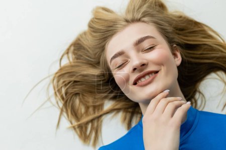 top view of happy blonde woman lying and touching chin on grey background 