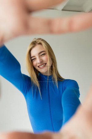 cheerful woman in blue turtleneck smiling at camera near blurred outstretched hands on grey background 