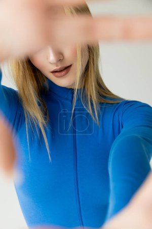 blonde woman in blue zipped turtleneck obscuring face with blurred hand isolated on grey