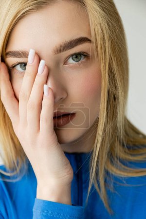 portrait of young woman with perfect skin and natural makeup holding hand on face isolated on grey