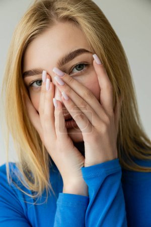 Photo for Portrait of blonde woman in blue long sleeve shirt looking at camera and covering face with hands isolated on grey - Royalty Free Image