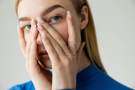 young blonde woman in blue turtleneck obscuring face with hands and looking at camera isolated on grey