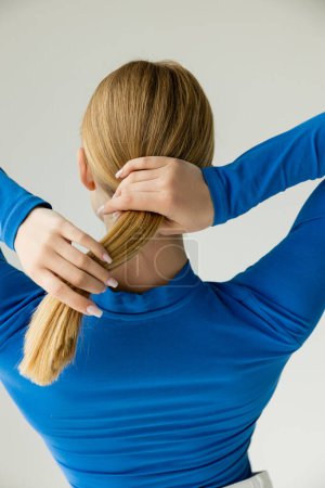 back view of blonde woman in blue turtleneck making hairstyle with ponytail isolated on grey