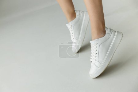 Foto de Vista recortada de la mujer en zapatillas de deporte de cuero blanco de pie en puntillas sobre fondo gris - Imagen libre de derechos