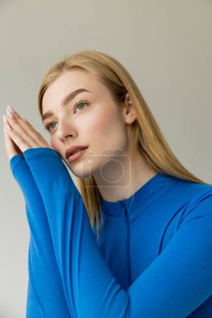 dreamy woman in blue long sleeve shirt holding hands near face and looking away isolated on grey