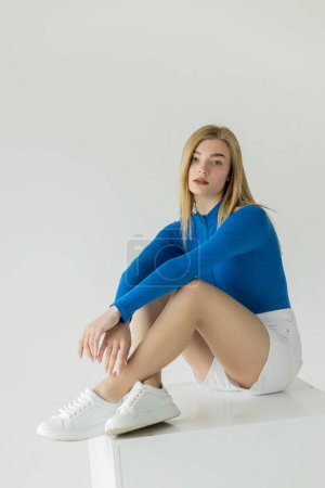 Téléchargez les photos : Jolie femme en chemise bleue à manches longues et baskets assises sur cube blanc et regardant la caméra isolée sur gris - en image libre de droit
