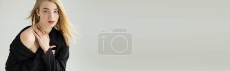 Téléchargez les photos : Séduisante femme blonde en veste noire regardant la caméra et touchant épaule nue isolé sur gris, bannière - en image libre de droit