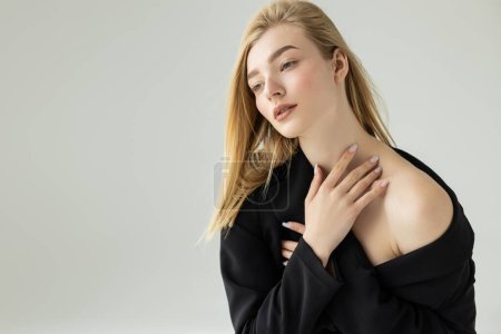 pretty and blonde woman in black jacket with bare shoulder touching neck isolated on grey