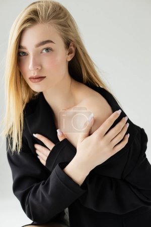 sensual woman with blonde hair and naked shoulder posing in black jacket and looking at camera isolated on grey