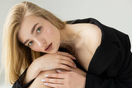 Photo for Sexy woman in black jacket embracing knee and looking at camera isolated on grey - Royalty Free Image