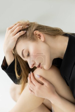 Photo for Side view of sexy blonde woman embracing knee and touching hair on grey background - Royalty Free Image