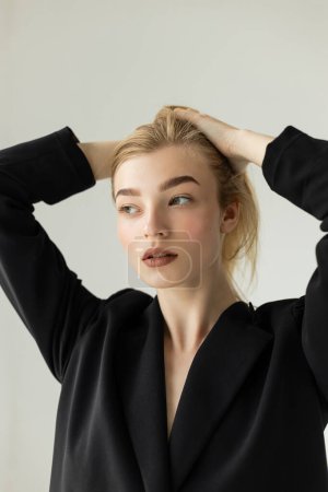 Téléchargez les photos : Femme blonde élégante en blazer noir ajustant les cheveux et regardant loin isolé sur gris - en image libre de droit
