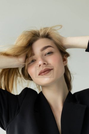 retrato de mujer rubia positiva con maquillaje natural ajustando el cabello y sonriendo a la cámara aislada en gris