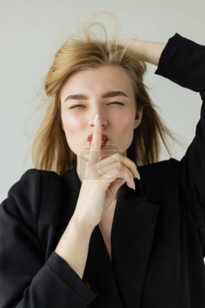 tricky woman in black blazer touching blonde hair and showing hush gesture isolated on grey