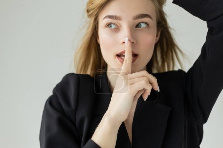 cunning woman in black blazer showing hush gesture and looking away isolated on grey