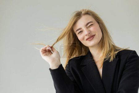 cheerful blonde woman in black jacket pulling hair and winking at camera isolated on grey