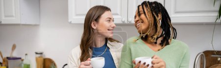 cheerful multiethnic lesbian couple holding cups of coffee and looking at each other in kitchen, banner