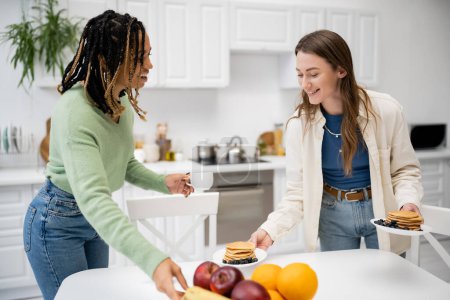 happy lesbian woman holding plates with tasty pancakes near african american girlfriend with cup of coffee