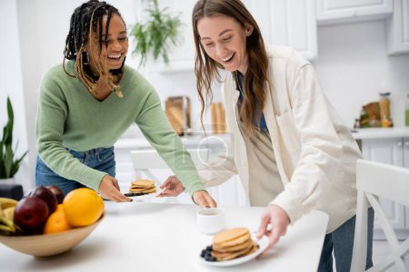 cheerful lesbian woman serving plates with tasty pancakes near african american girlfriend 