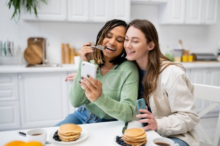 happy african american lesbian woman showing smartphone to girlfriend during breakfast 