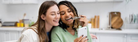 happy african american lesbian woman showing smartphone to girlfriend in kitchen, banner