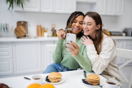 cheerful african american lesbian woman showing smartphone to happy girlfriend during breakfast 