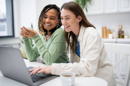 fröhliche lesbische Frau mit Laptop in der Nähe lächelnde afrikanisch-amerikanische Freundin hält Tasse Kaffee 