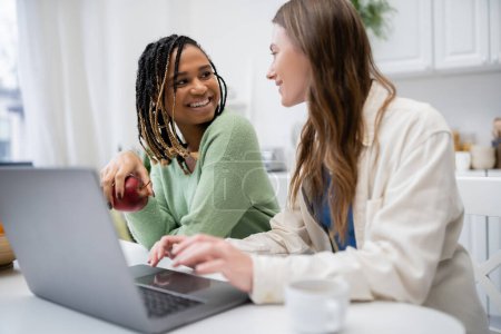 cheerful lesbian woman using laptop near smiling african american girlfriend holding apple 