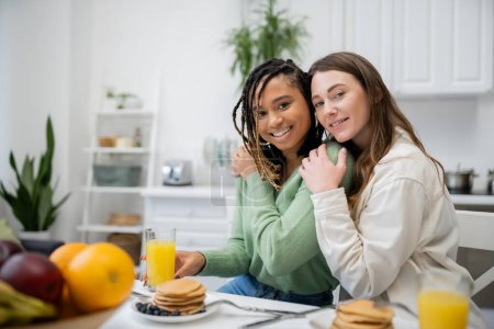 fröhliche lesbische Frau umarmt lächelnde afrikanisch-amerikanische Freundin beim Frühstück 