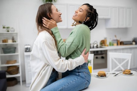 freudige und lesbische afrikanisch-amerikanische Frau sitzt am Esstisch und umarmt glückliche Freundin