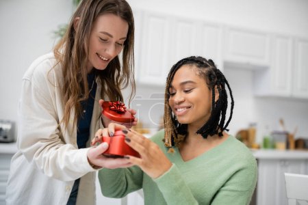 Téléchargez les photos : Heureuse lesbienne femme tenant en forme de coeur boîte cadeau près souriant afro-américaine copine - en image libre de droit