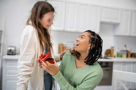Téléchargez les photos : Heureuse femme lesbienne tenant boîte cadeau en forme de coeur près étonnante petite amie afro-américaine - en image libre de droit
