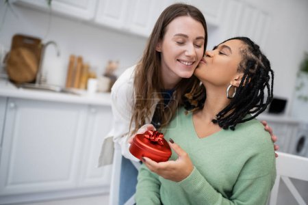 Foto de Mujer lesbiana afroamericana besándose mejilla de novia alegre y sosteniendo caja de regalo en forma de corazón - Imagen libre de derechos