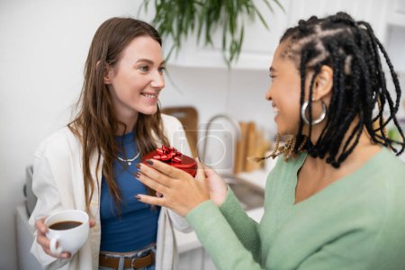 Téléchargez les photos : Africaine américaine lesbienne femme tenant boîte cadeau rouge près petite amie heureuse avec tasse de café le jour de la Saint-Valentin - en image libre de droit