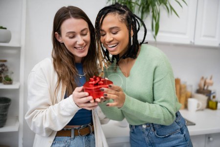 Téléchargez les photos : Heureux interracial et lesbiennes femmes tenant rouge boîte cadeau en forme de coeur avec arc sur la Saint-Valentin - en image libre de droit