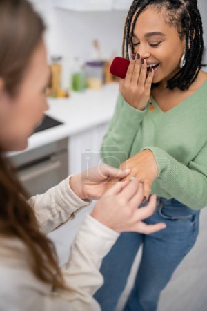 lesbian woman making proposal and wearing engagement ring on finger of amazed african american girlfriend