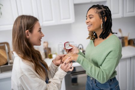 lesbian woman making proposal and wearing engagement ring on finger of happy african american girlfriend on valentines day